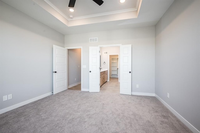 unfurnished bedroom featuring light carpet, a tray ceiling, ornamental molding, ensuite bathroom, and ceiling fan