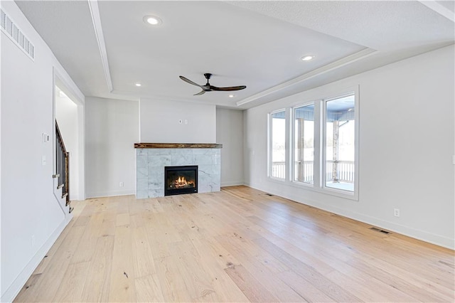 unfurnished living room with a raised ceiling, a tile fireplace, ceiling fan, and light wood-type flooring