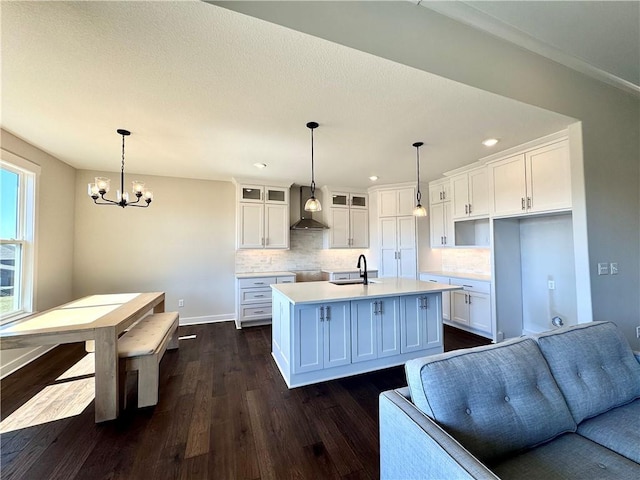 kitchen with dark wood-type flooring, a center island with sink, a sink, white cabinetry, and wall chimney range hood