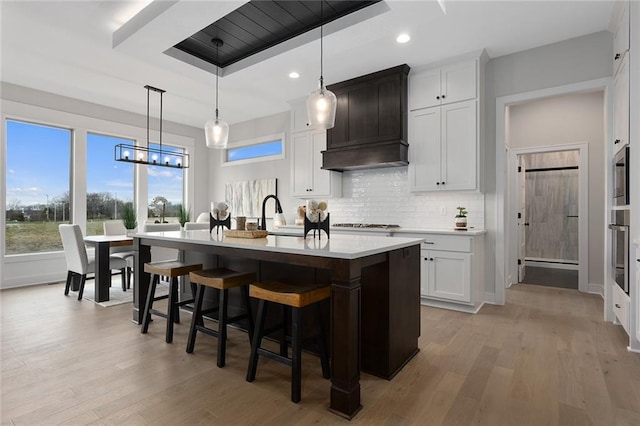 kitchen with light hardwood / wood-style floors, decorative light fixtures, a center island with sink, white cabinets, and custom exhaust hood