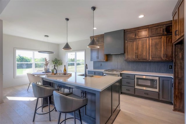 kitchen with pendant lighting, an island with sink, light hardwood / wood-style flooring, wall chimney range hood, and appliances with stainless steel finishes