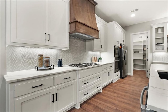 kitchen with hardwood / wood-style floors, custom exhaust hood, stainless steel appliances, white cabinets, and light stone counters