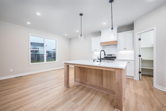 kitchen featuring stainless steel range, custom range hood, a kitchen island with sink, pendant lighting, and white cabinetry