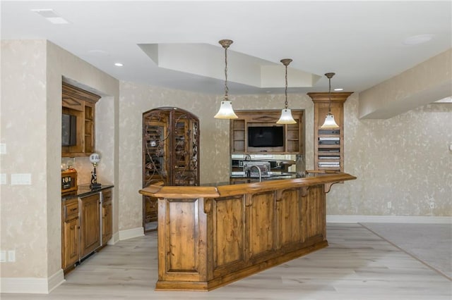 kitchen featuring a tray ceiling, brown cabinets, a center island with sink, light wood-style floors, and wallpapered walls