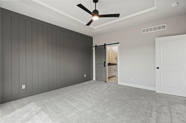 unfurnished bedroom with a barn door, connected bathroom, a tray ceiling, ceiling fan, and light colored carpet