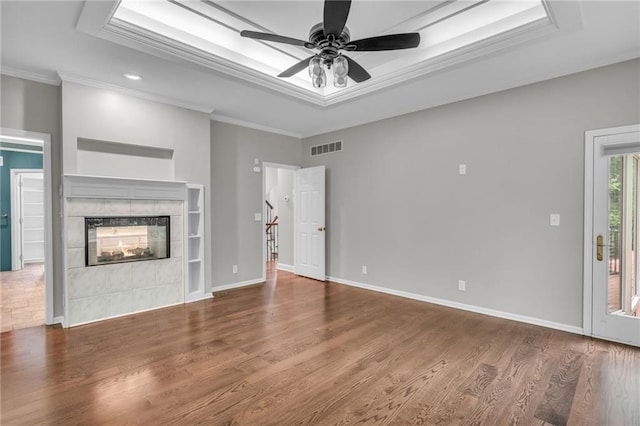 unfurnished living room with ceiling fan, wood-type flooring, a tray ceiling, a tiled fireplace, and ornamental molding