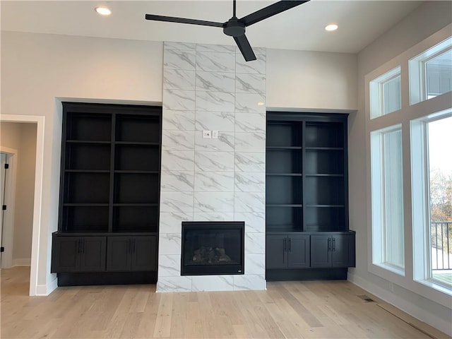 unfurnished living room featuring a tiled fireplace, built in shelves, ceiling fan, and light wood-type flooring