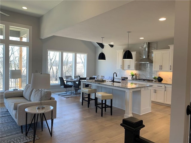 kitchen featuring wall chimney range hood, a breakfast bar, a kitchen island with sink, light stone counters, and white cabinets