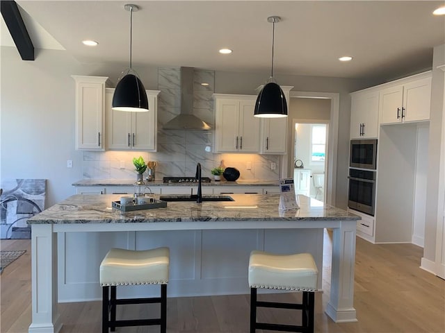 kitchen featuring a kitchen island with sink, light stone counters, white cabinetry, and wall chimney exhaust hood