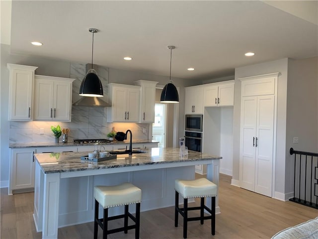 kitchen featuring appliances with stainless steel finishes, an island with sink, sink, dark stone countertops, and wall chimney range hood