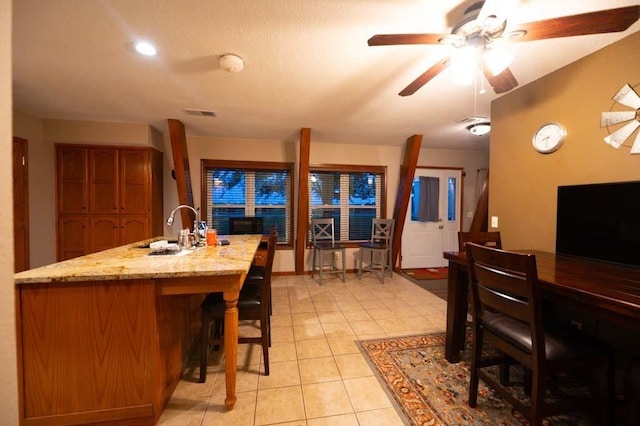 kitchen with sink, ceiling fan, an island with sink, light tile patterned flooring, and light stone counters