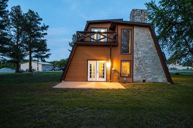 back house at dusk with a lawn, a balcony, a patio, and french doors