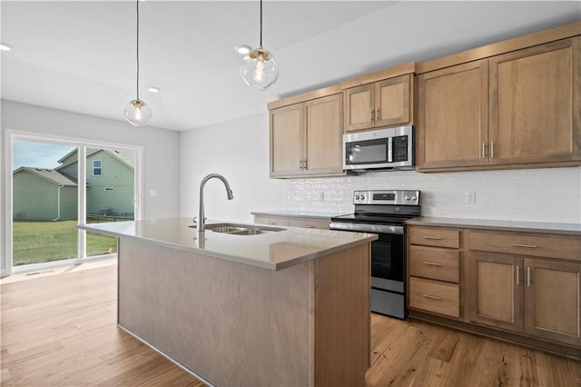 kitchen with light wood-type flooring, stainless steel appliances, sink, pendant lighting, and an island with sink