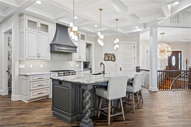 kitchen with custom range hood, hanging light fixtures, glass insert cabinets, a sink, and an island with sink