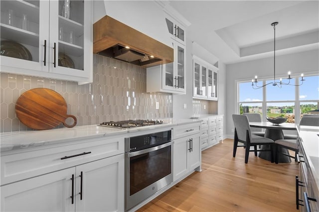 kitchen featuring light wood-style flooring, stainless steel appliances, custom exhaust hood, light stone countertops, and an inviting chandelier