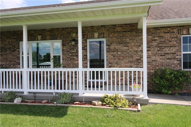 view of exterior entry with covered porch, brick siding, a lawn, and a shingled roof