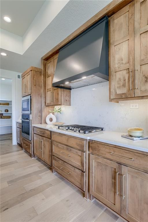 kitchen with stainless steel oven, wall chimney range hood, light countertops, and light wood-type flooring