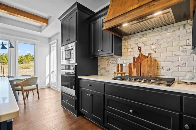 kitchen with appliances with stainless steel finishes, dark hardwood / wood-style flooring, beamed ceiling, tasteful backsplash, and premium range hood