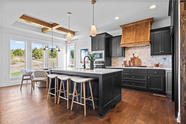 kitchen featuring custom range hood, decorative light fixtures, an island with sink, coffered ceiling, and beamed ceiling