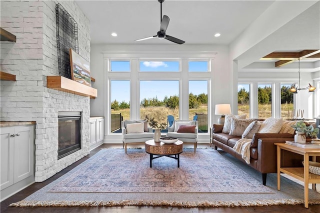 living room with ceiling fan with notable chandelier, a brick fireplace, beam ceiling, and dark hardwood / wood-style flooring