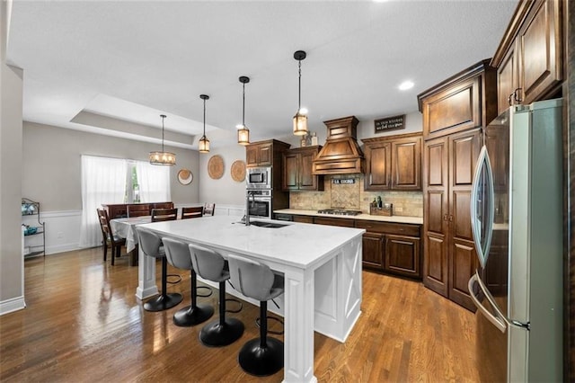 kitchen featuring premium range hood, a breakfast bar area, hanging light fixtures, appliances with stainless steel finishes, and an island with sink