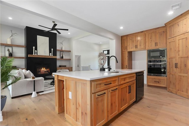 kitchen with a large fireplace, sink, an island with sink, light hardwood / wood-style floors, and black appliances