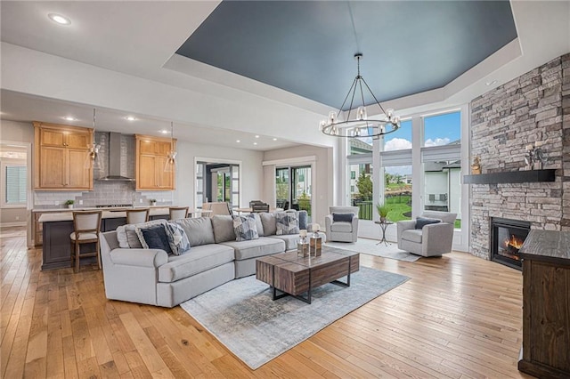 living room featuring an inviting chandelier, a stone fireplace, a raised ceiling, and light wood-type flooring