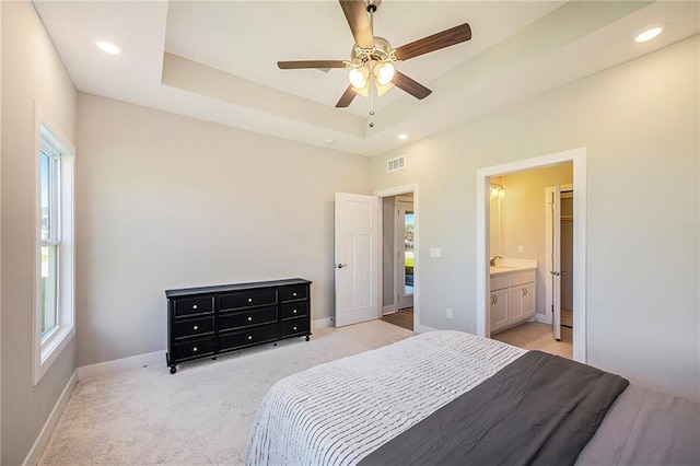 bedroom featuring ceiling fan, sink, ensuite bathroom, light colored carpet, and a tray ceiling