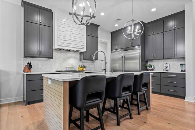 kitchen with an island with sink, stainless steel built in refrigerator, light wood-type flooring, and a kitchen bar