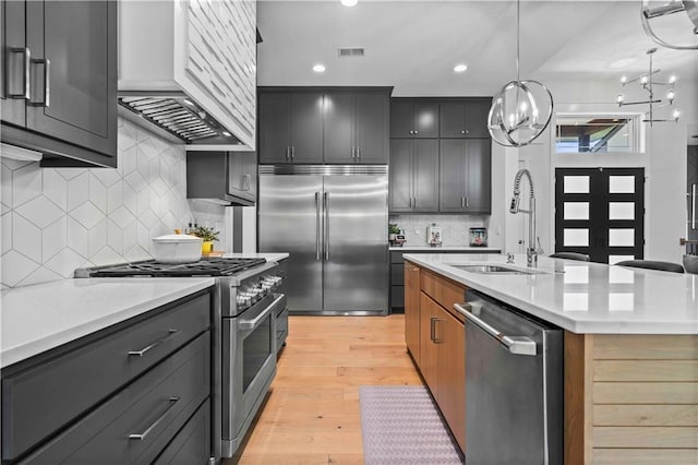 kitchen featuring a kitchen island with sink, hanging light fixtures, high end appliances, sink, and light hardwood / wood-style flooring