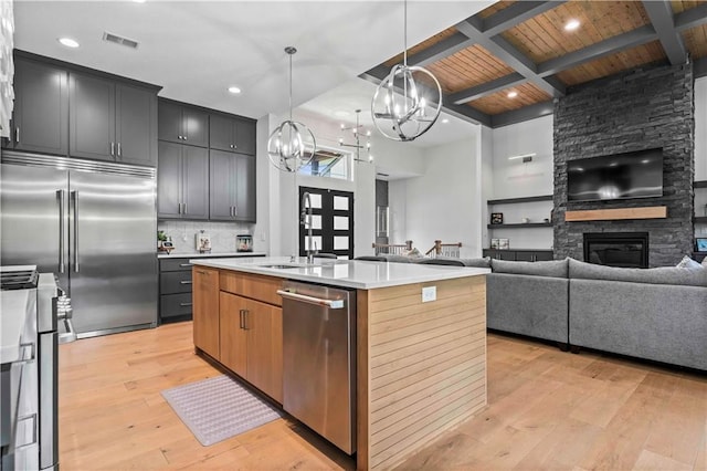 kitchen with a stone fireplace, appliances with stainless steel finishes, hanging light fixtures, an island with sink, and beam ceiling