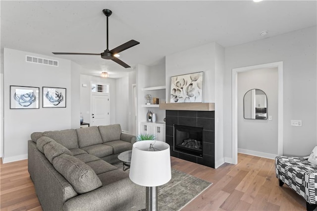 living room with built in features, light wood-type flooring, a tile fireplace, and ceiling fan