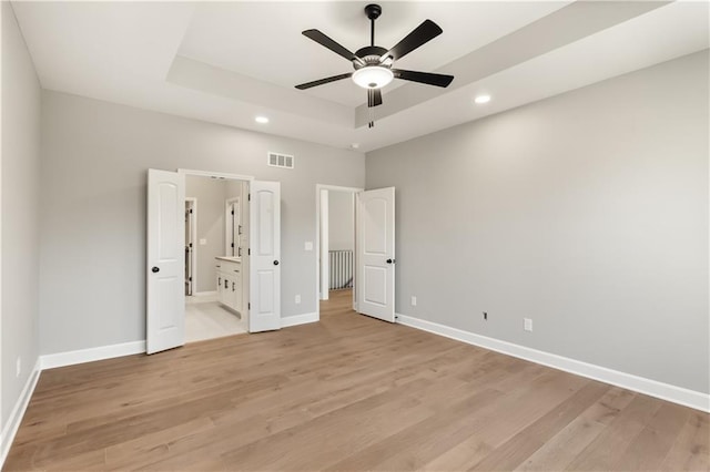 unfurnished bedroom featuring ensuite bathroom, a raised ceiling, ceiling fan, and light hardwood / wood-style floors