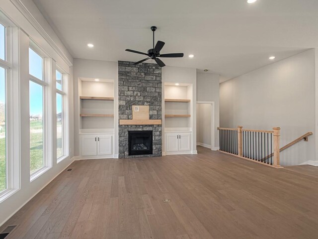 unfurnished living room with ceiling fan, a stone fireplace, built in shelves, and hardwood / wood-style floors