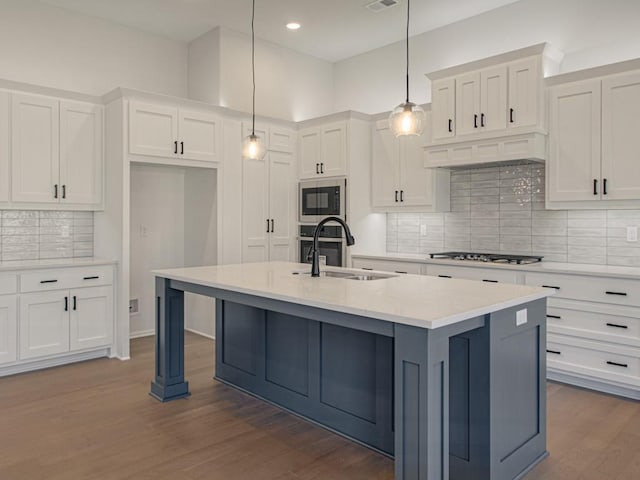 kitchen featuring appliances with stainless steel finishes, hanging light fixtures, white cabinets, hardwood / wood-style flooring, and a center island with sink