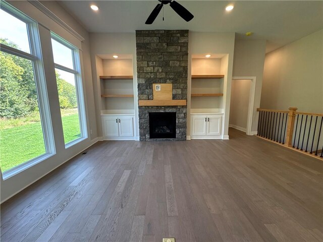 unfurnished living room featuring ceiling fan, a stone fireplace, built in shelves, and wood-type flooring