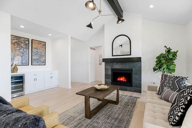 living room featuring vaulted ceiling with beams, beverage cooler, a tile fireplace, and light wood-type flooring