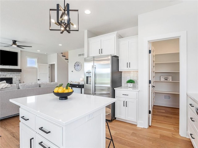 kitchen with white cabinetry, stainless steel refrigerator with ice dispenser, a fireplace, a kitchen island, and ceiling fan with notable chandelier