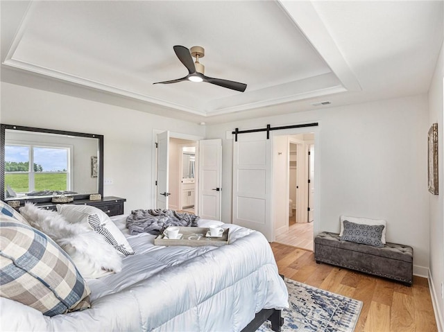 bedroom with a barn door, ceiling fan, a tray ceiling, ensuite bath, and light wood-type flooring