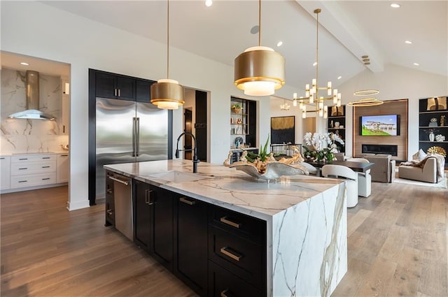 kitchen with sink, light stone counters, decorative light fixtures, a kitchen island with sink, and exhaust hood