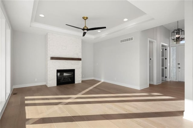 unfurnished living room with ceiling fan with notable chandelier, wood-type flooring, a fireplace, and a tray ceiling