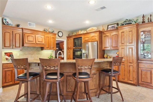 kitchen featuring decorative backsplash, light stone counters, an island with sink, and black appliances