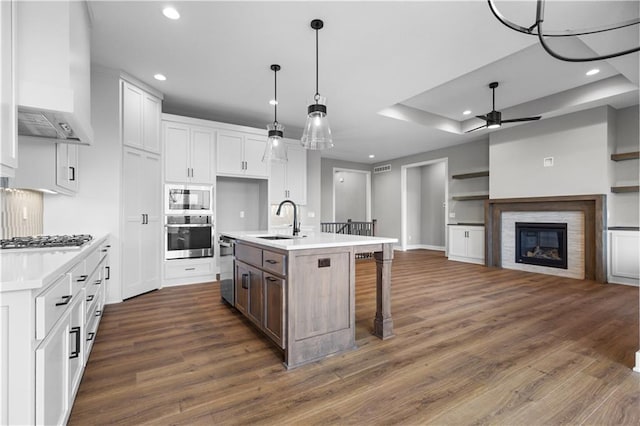 kitchen with wall chimney exhaust hood, stainless steel appliances, sink, a center island with sink, and white cabinetry