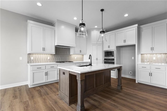 kitchen featuring custom exhaust hood, built in microwave, sink, pendant lighting, and an island with sink