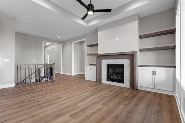 unfurnished living room featuring a raised ceiling, built in shelves, ceiling fan, and light hardwood / wood-style floors