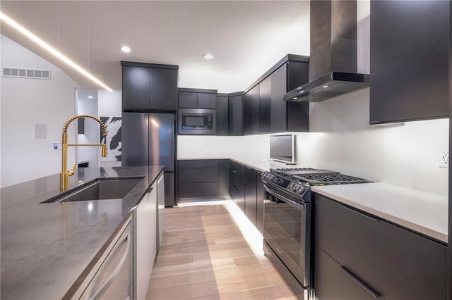 kitchen with wall chimney range hood, sink, light hardwood / wood-style flooring, appliances with stainless steel finishes, and dark stone counters