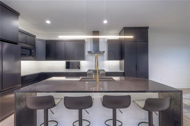 kitchen featuring sink, a breakfast bar area, a kitchen island with sink, built in appliances, and wall chimney range hood