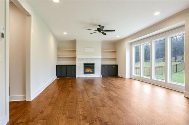 unfurnished living room featuring built in shelves, ceiling fan, dark wood-type flooring, crown molding, and a tiled fireplace