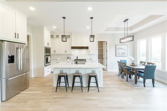 kitchen featuring appliances with stainless steel finishes, decorative light fixtures, a kitchen island with sink, white cabinets, and light wood-type flooring