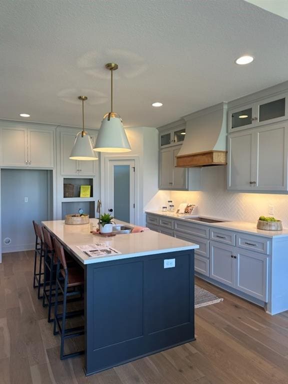 kitchen with dark wood-type flooring, an island with sink, a sink, black electric cooktop, and custom exhaust hood